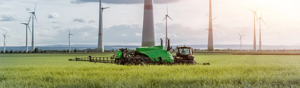 Traktor ute i fält. I bakgrunden syns vindkraftverk.